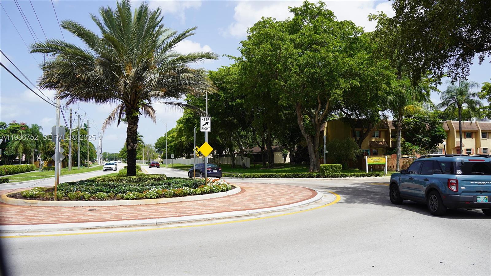 A blue SUV drives past a landscaped roundabout with a palm tree and traffic sign — 7175 Nova Dr # 204, Davie FL 33317, DAVIE, FL