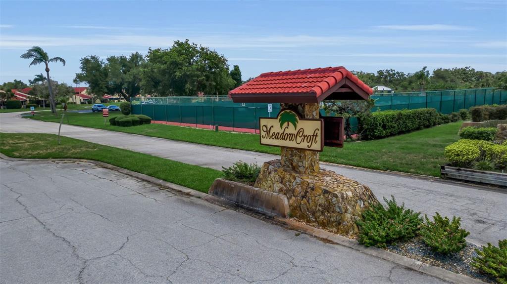 A stone monument sign for Meadow Croft stands near a tennis court and paved road — 1409 56TH STREET W UNIT 1409, BRADENTON, FL
