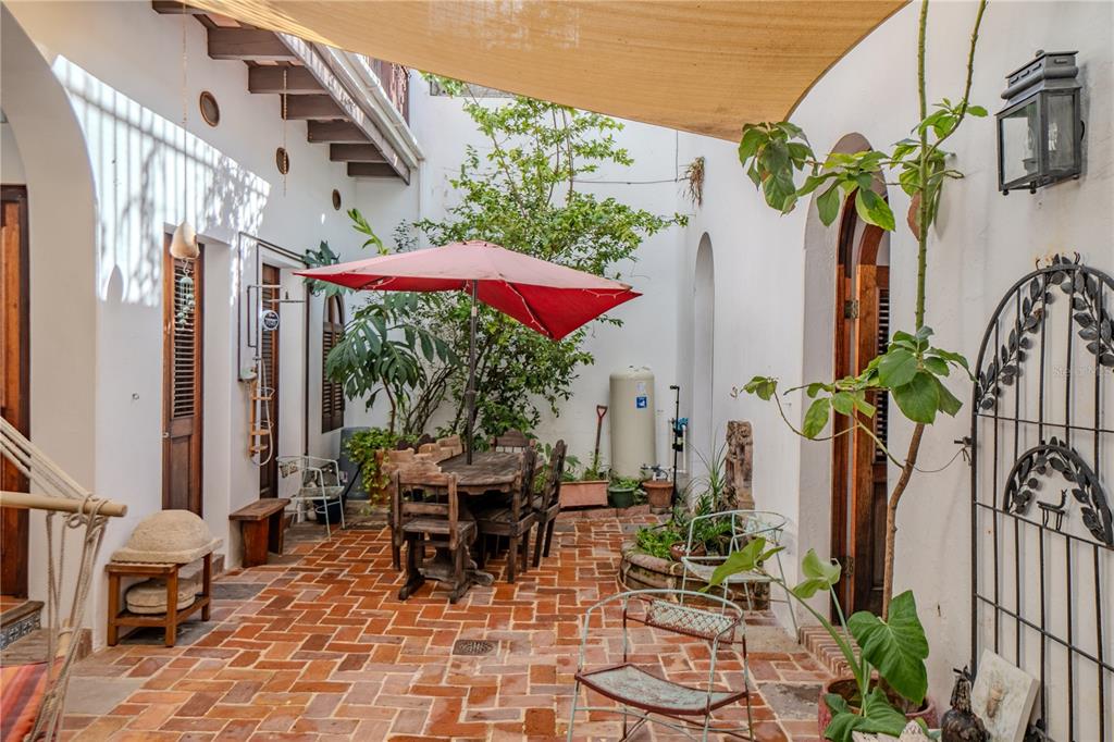 An outdoor courtyard features brick flooring, a dining set under a red umbrella, and plants — 3 CRUZ, SAN JUAN, PR