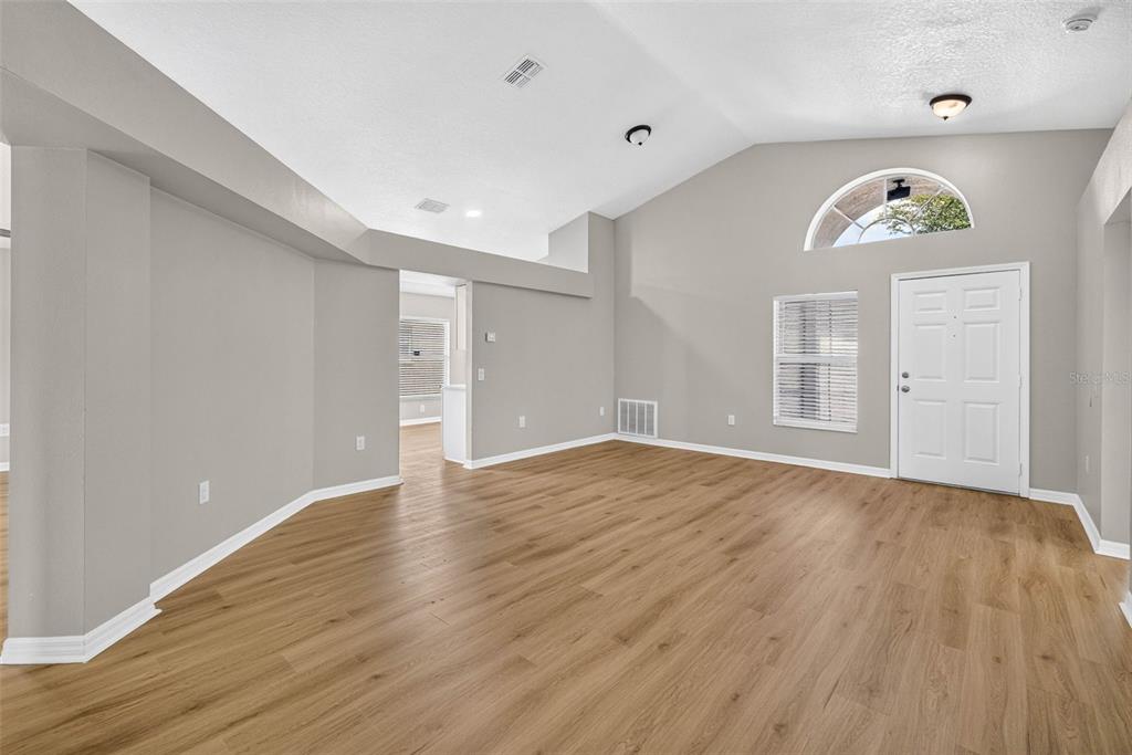 An empty living room features light wood-look flooring, vaulted ceilings, and a white front door — 25442 BRUFORD BOULEVARD, LAND O LAKES, FL