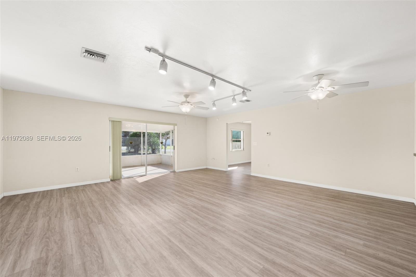 A spacious, empty living room features light wood-look flooring, ceiling fans, and track lighting — 1273 Woodbury Avenue, Sebring FL 33875, SEBRING, FL