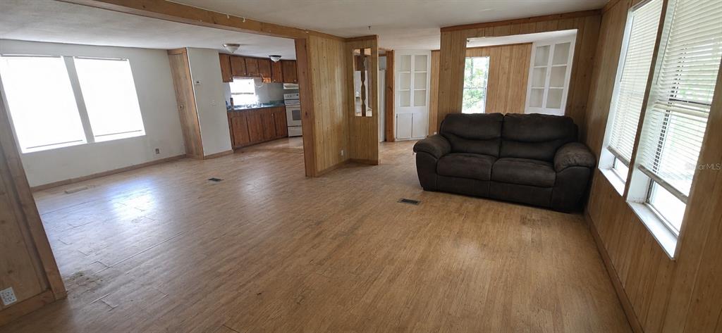 A living room with wood-paneled walls and light wood flooring leads into a kitchen area — 702 DAVID AVENUE, INTERLACHEN, FL