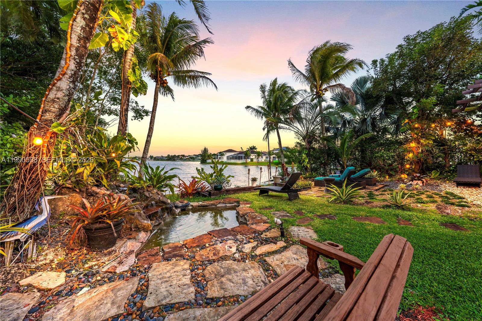 A backyard garden features a stone-lined pond, lounge chairs, and palm trees overlooking a lake — 5265 SW 171st Ave, Miramar FL 33027, MIRAMAR, FL