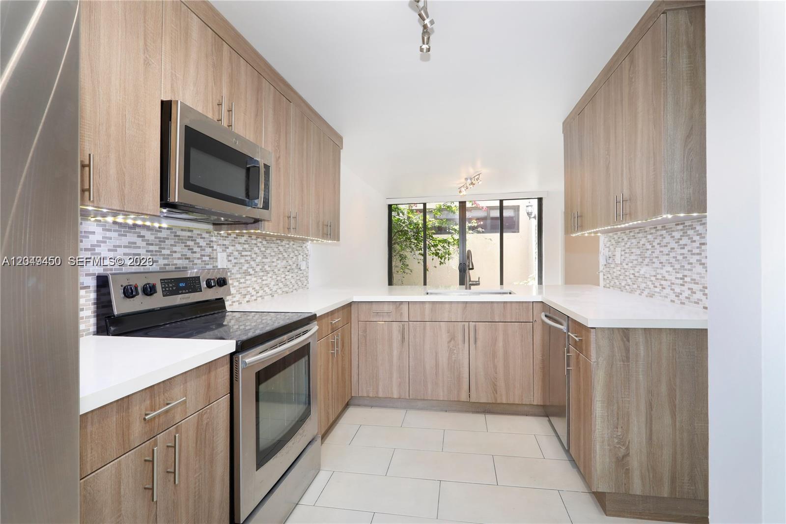 A modern kitchen features wood-grain cabinets, stainless steel appliances, white countertops, and a tiled backsplash — 8124 SW 87th Ter, Miami FL 33143, MIAMI, FL