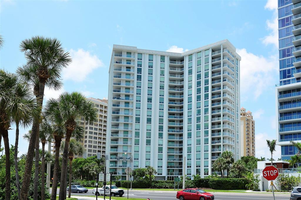 A tall white residential high-rise building stands behind palm trees and a street with cars — 1111 N GULFSTREAM AVENUE UNIT PH-C, SARASOTA, FL