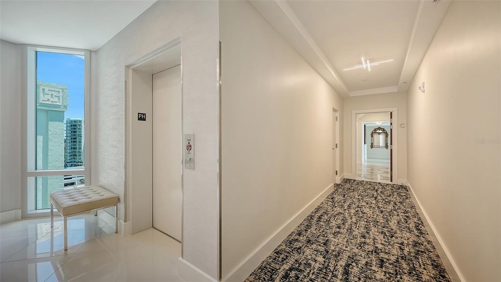 A hallway features a white elevator, a patterned carpet, and a window with city views — 1111 N GULFSTREAM AVENUE UNIT PH-C, SARASOTA, FL