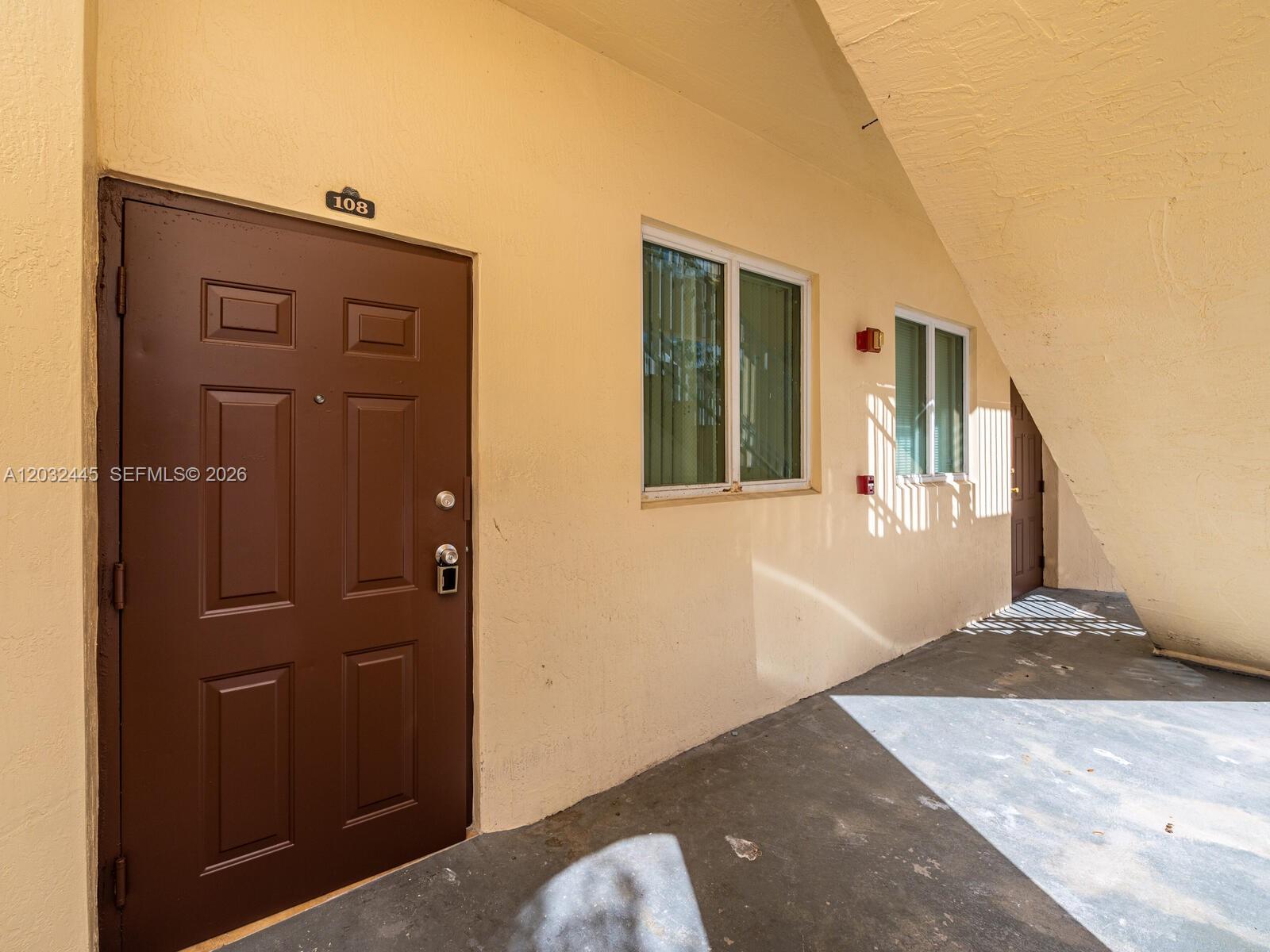 A brown door labeled 108 sits next to windows on a beige exterior walkway — 1663 SE 27th Dr # 108, Homestead FL 33035, HOMESTEAD, FL