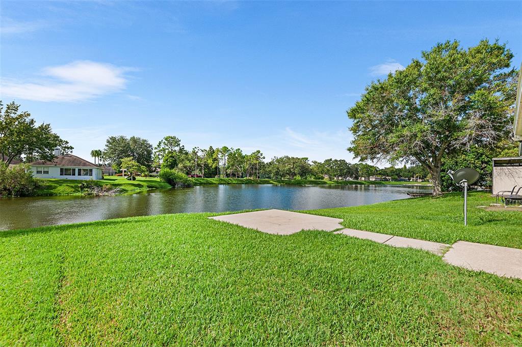 A grassy backyard features a concrete patio overlooking a pond with trees in the distance — 2101 HATTERAS POINT, LAKELAND, FL