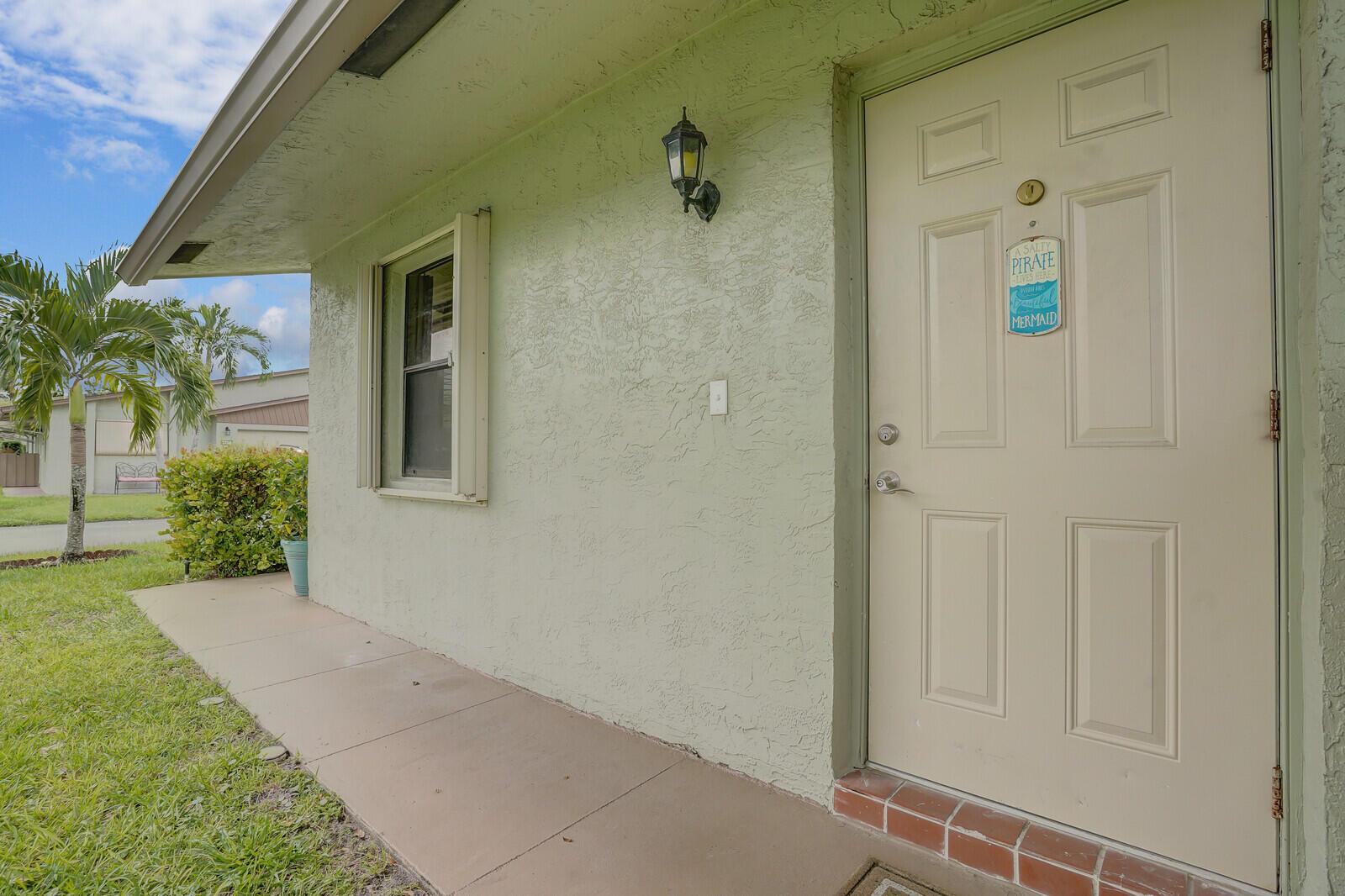 A light green stucco exterior wall features a front door with a decorative sign — 1737 Palmland Drive, Boynton Beach FL 33436, BOYNTON BEACH, FL