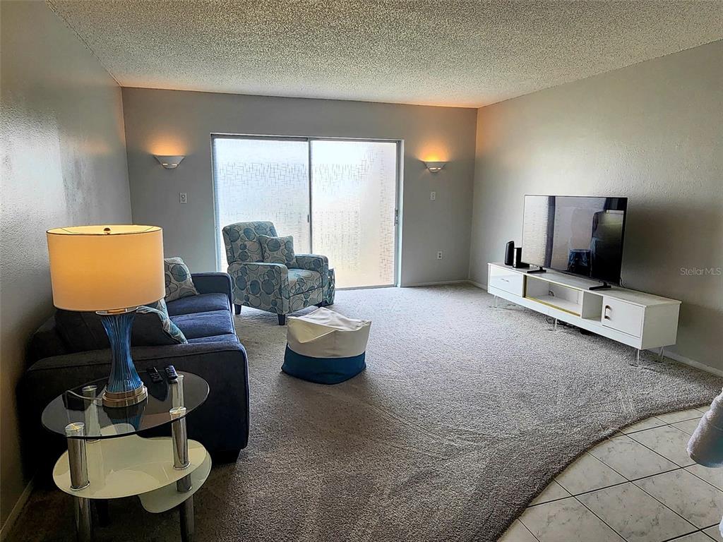 A living room features gray carpet, a blue sofa, a patterned armchair, and a television console — 4215 E BAY DRIVE UNIT 1602D, CLEARWATER, FL
