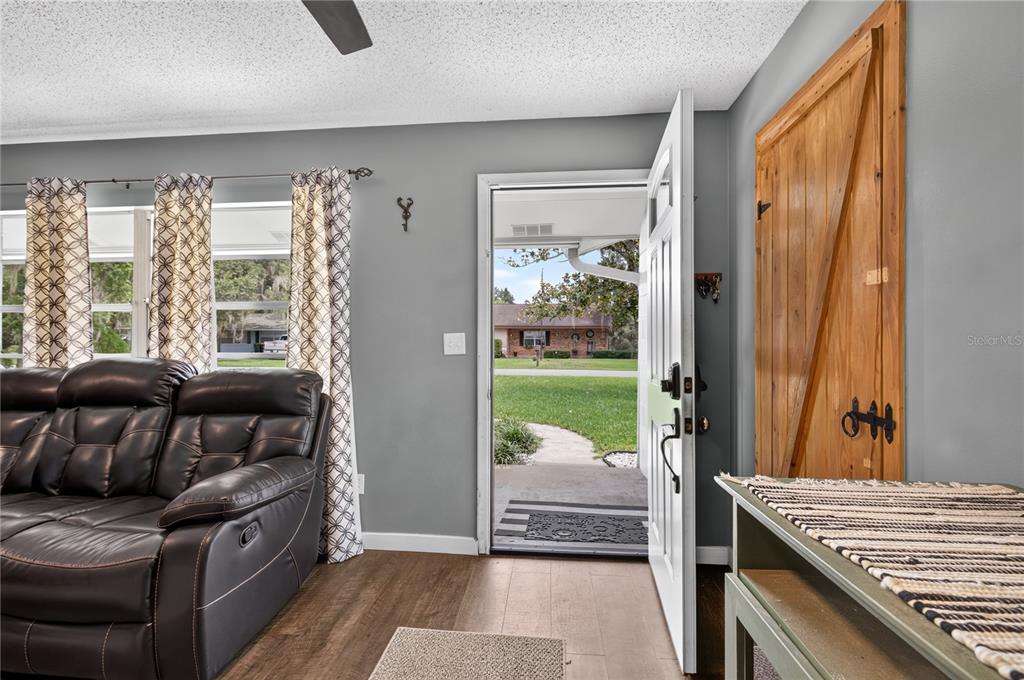 A living room with a dark leather sofa, wood-look flooring, and an open front door — 555 WINOGENE AVE, UMATILLA, FL