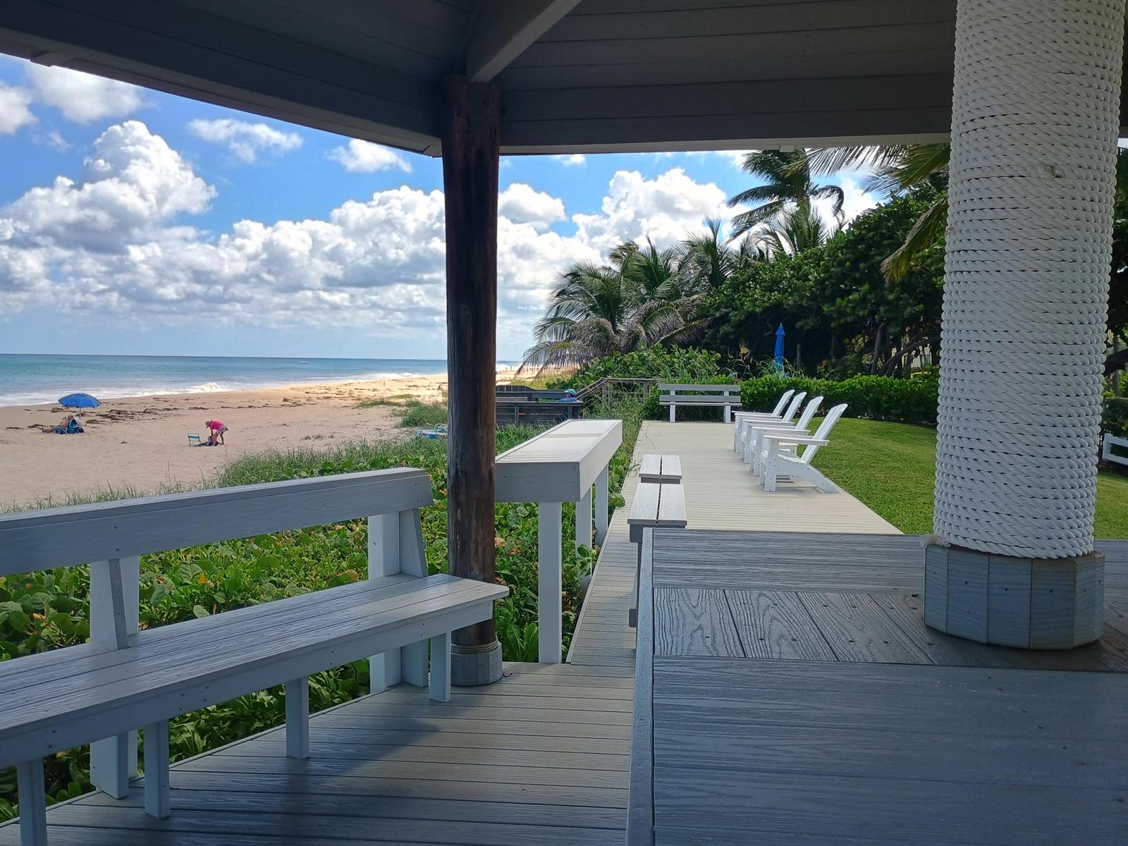 A covered wooden deck overlooks a sandy beach with white benches and chairs — 5505 N Ocean Boulevard # 106, Ocean Ridge FL 33435, OCEAN RIDGE, FL