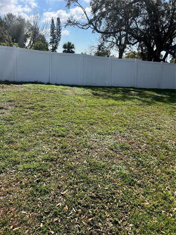 A grassy backyard features a white vinyl privacy fence and mature trees under a blue sky — 7438 COVENTRY DRIVE, PORT RICHEY, FL