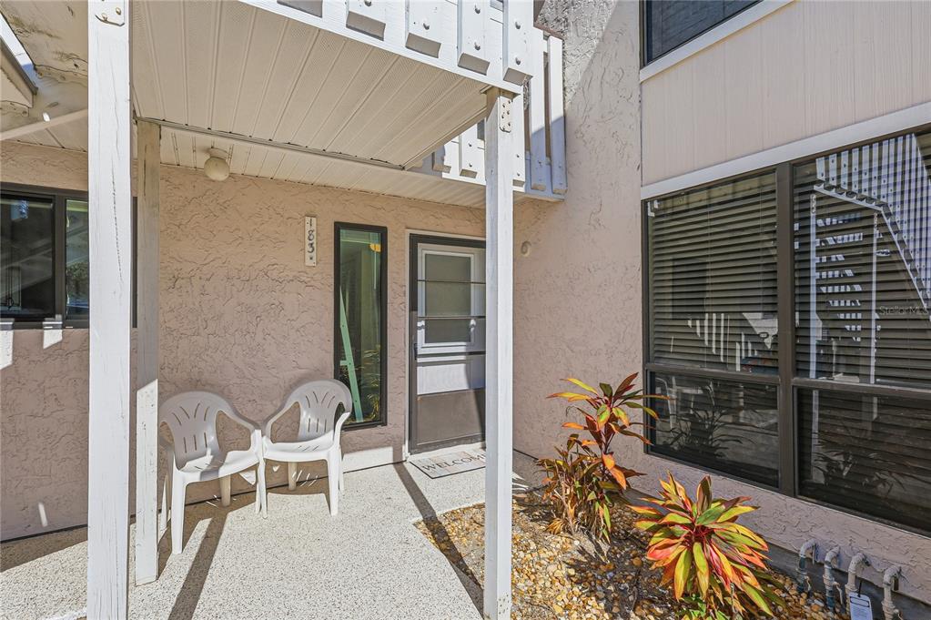A covered entryway with two white plastic chairs, a front door, and small landscaping plants — 1801 GULF DRIVE N UNIT 183, BRADENTON BEACH, FL