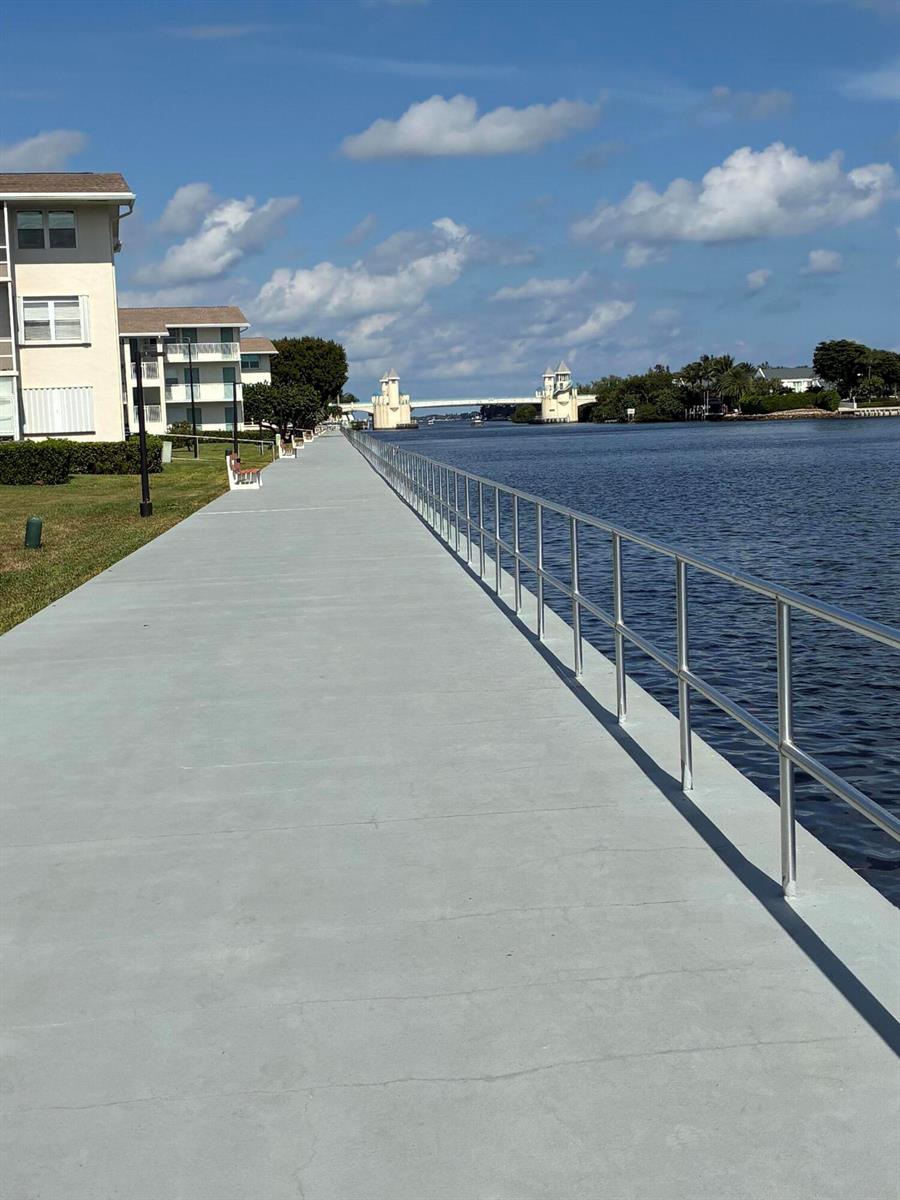 A concrete walkway with a metal railing runs alongside a canal toward a distant bridge — 650 Horizons Avenue E # 107, Boynton Beach FL 33435, BOYNTON BEACH, FL