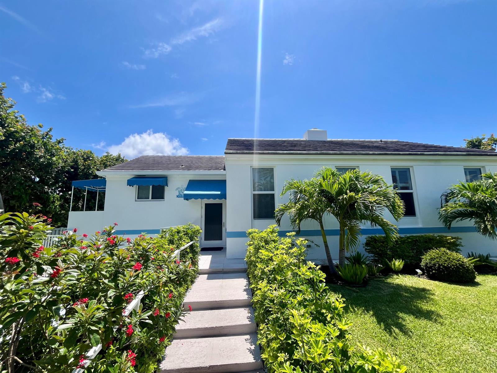 A white house with blue awnings and a palm tree sits behind a landscaped walkway — 115 Dolphin Road, Ocean Ridge FL 33435, OCEAN RIDGE, FL