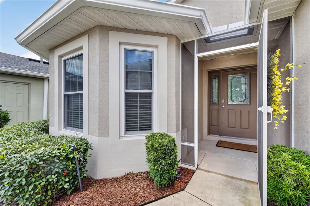 A tan house exterior features a screened entryway, a brown front door, and two windows — 27134 NOSTALGIA DRIVE, LEESBURG, FL