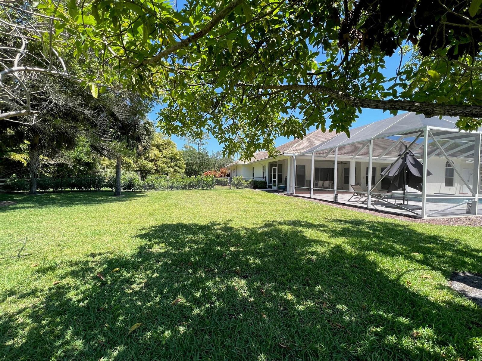 A grassy backyard features a screened-in pool enclosure attached to a single-story house — 103 Squire Drive, Wellington FL 33414, WELLINGTON, FL