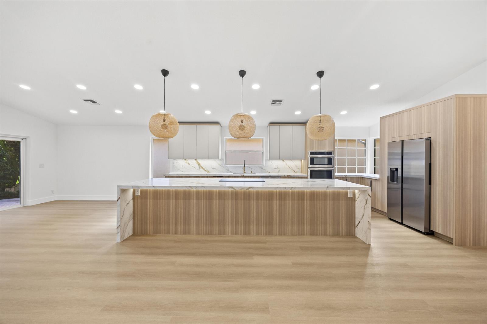 A modern kitchen features a large marble waterfall island, light wood cabinetry, and woven pendant lights — 5141 Columbo Court, Delray Beach FL 33484, DELRAY BEACH, FL