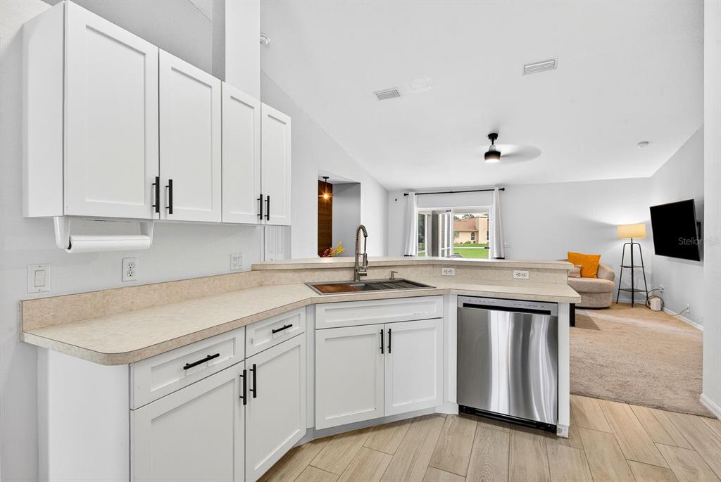 A kitchen with white cabinets, stainless steel dishwasher, and beige countertops overlooks a living area — 4394 CUTHBERT AVENUE, NORTH PORT, FL