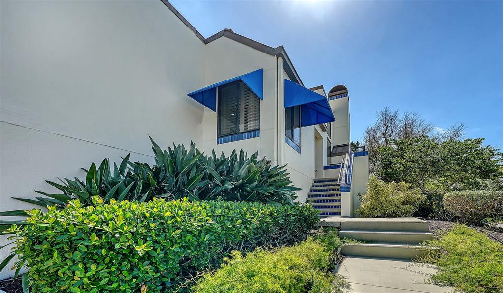 A white building exterior features blue window awnings, a tiled staircase, and lush green landscaping — 2000 HARBOURSIDE DRIVE UNIT 1501, LONGBOAT KEY, FL