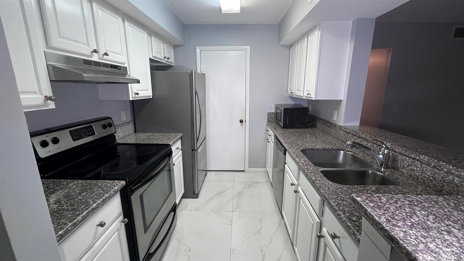 A galley kitchen features white cabinets, granite countertops, stainless steel appliances, and light-colored tile flooring — 815 W Boynton Beach Boulevard # 107, Boynton Beach FL 33426, BOYNTON BEACH, FL