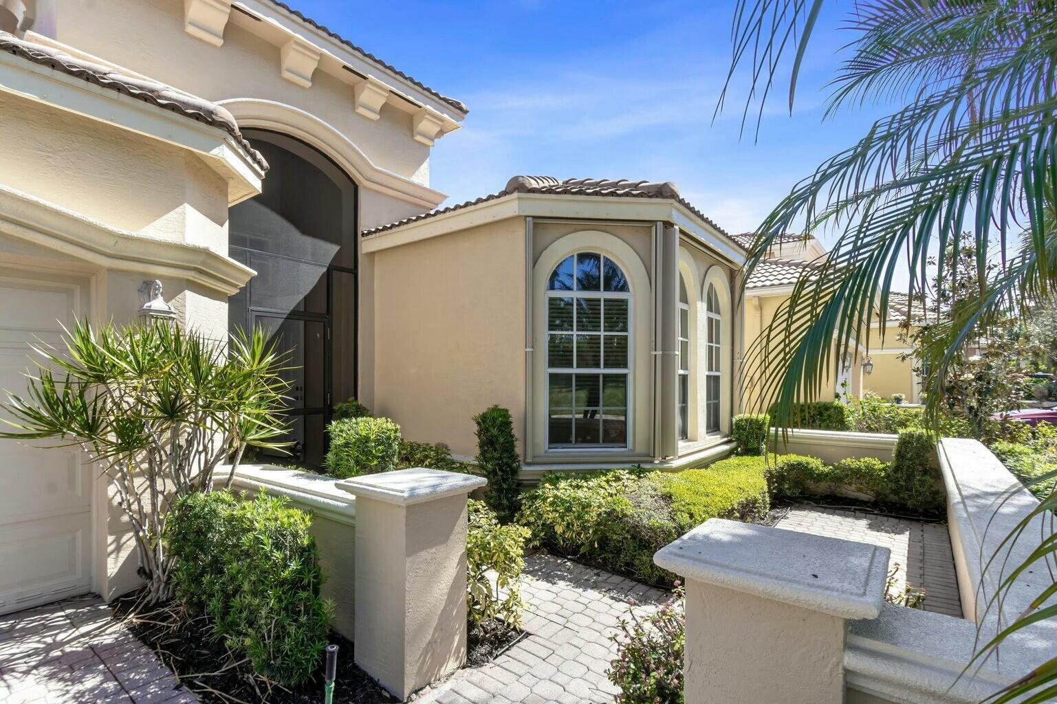A paved walkway leads to the entrance of a beige stucco house with arched windows — 8709 Via Grande E, Wellington FL 33411, WELLINGTON, FL