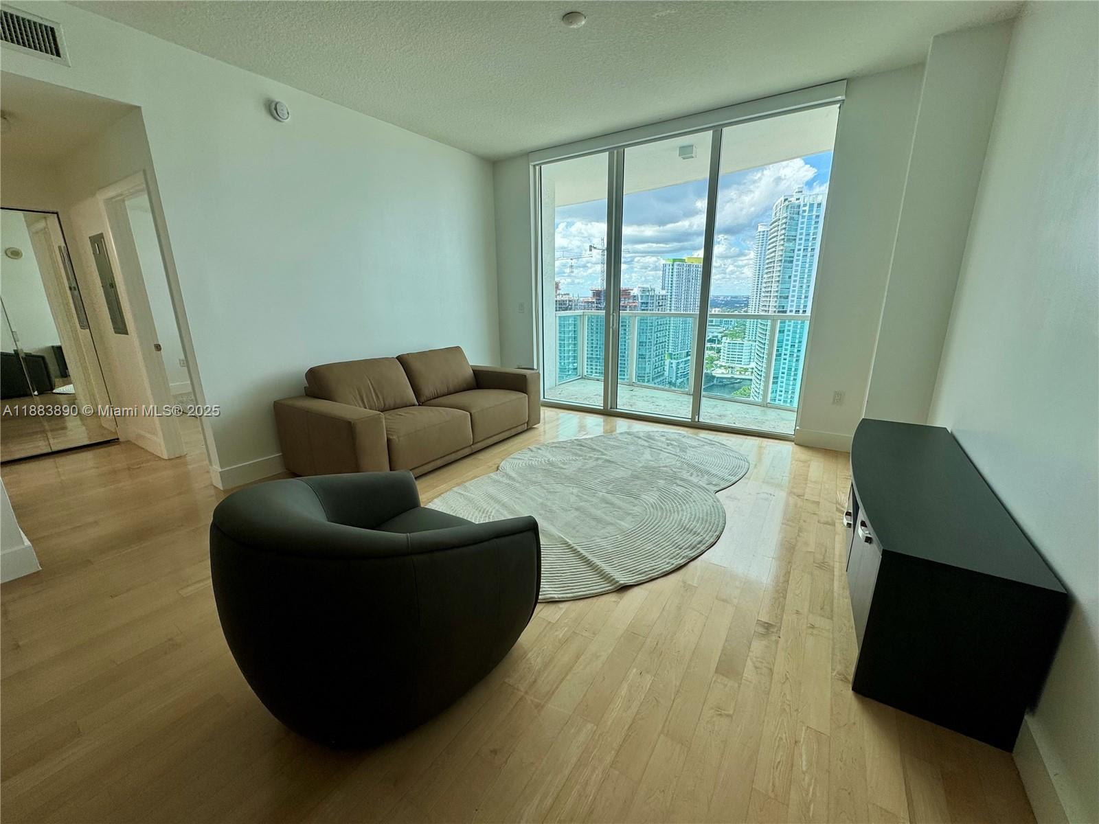 A living room features a brown sofa, a dark armchair, and floor-to-ceiling windows overlooking city buildings — 31 SE 5th St # 3817, Miami FL 33131, MIAMI, FL
