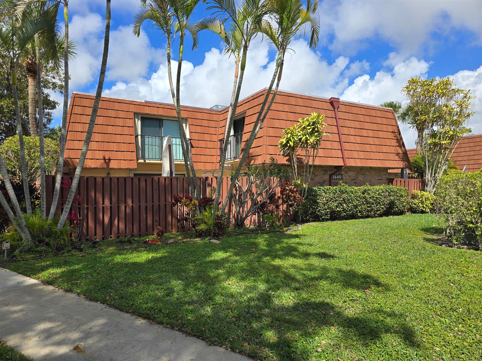 A two-story building with a brown mansard roof, wooden fence, and palm trees on a lawn — 1440 Augusta Circle # 132, Delray Beach FL 33445, DELRAY BEACH, FL