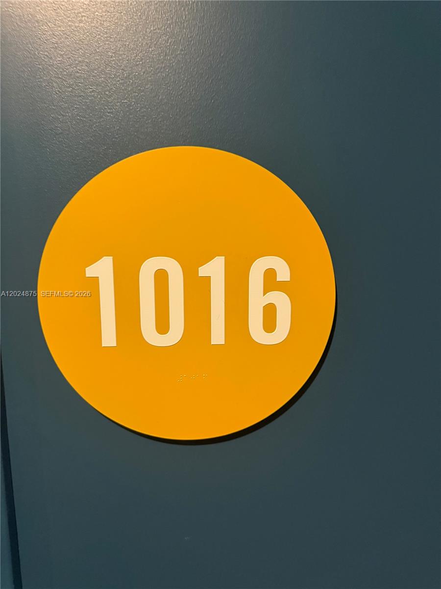 A yellow circular sign with the number 1016 and braille is mounted on a wall — 90 NE 32 # 1016, Miami FL 33137, MIAMI, FL