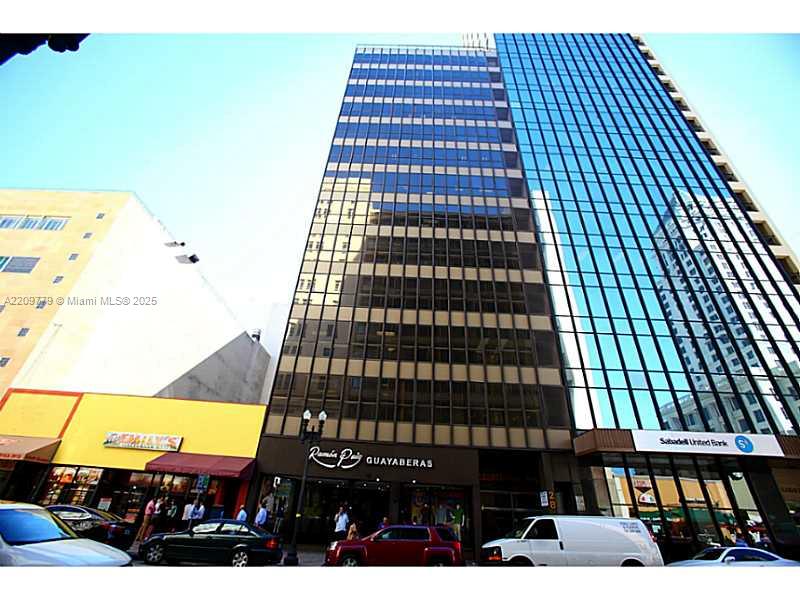 A tall office building with a glass facade stands above street-level retail shops and parking — 28 W FLAGLER ST, Miami FL 33130, MIAMI, FL