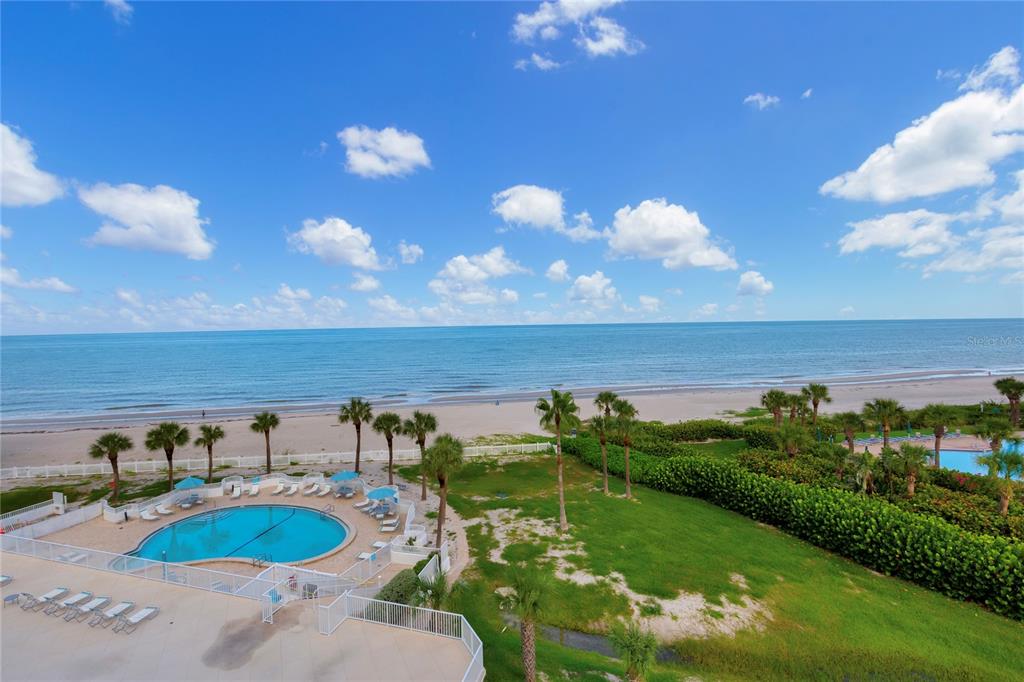 An elevated view of a swimming pool, grassy lawn, palm trees, and the ocean beach — 1582 GULF BOULEVARD UNIT 1506, CLEARWATER BEACH, FL