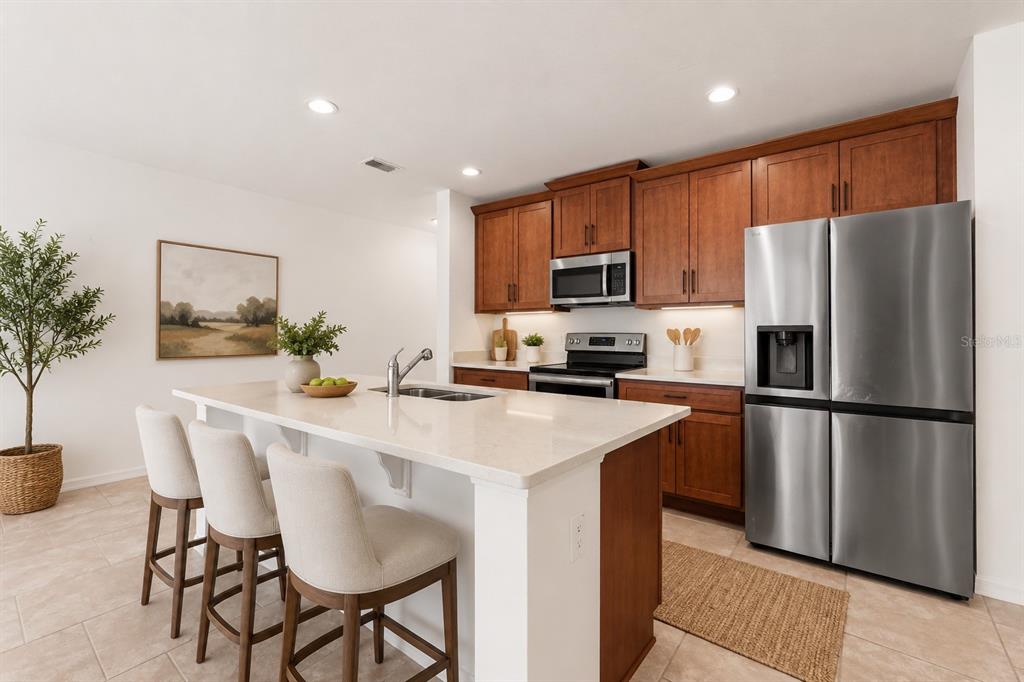 A kitchen features wood cabinets, stainless steel appliances, a quartz island, and three bar stools — 1259 WISPY CYPRESS DRIVE, KISSIMMEE, FL