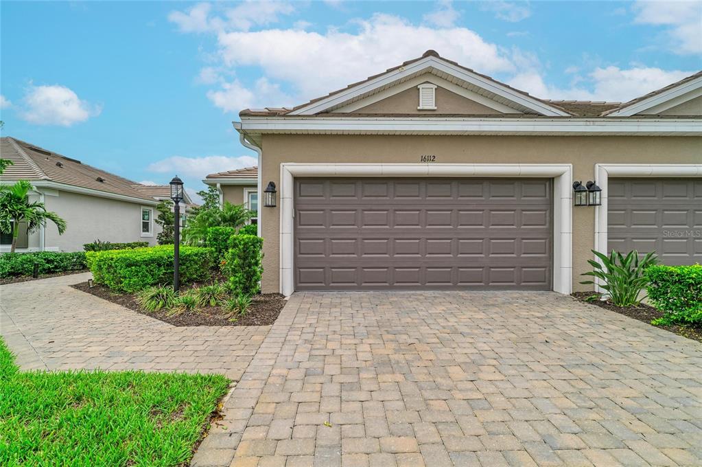 A beige house exterior features a two-car garage, a paved driveway, and landscaped front bushes — 16112 ISOLA PLACE, BRADENTON, FL
