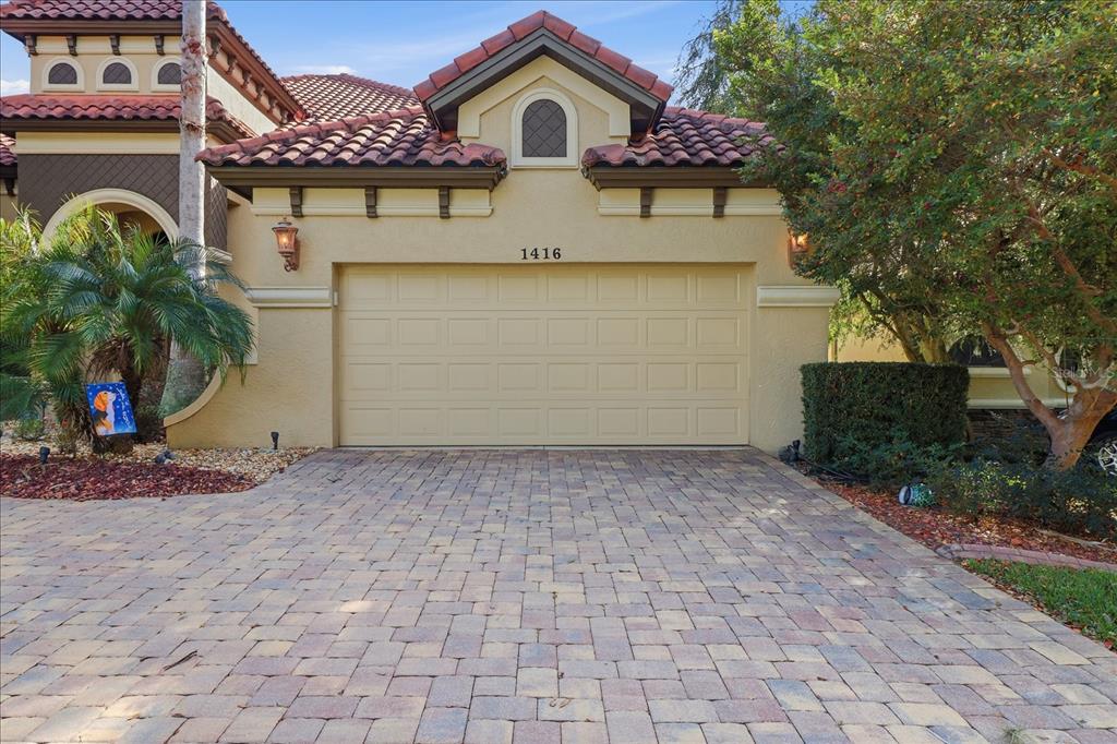 A beige house exterior features a two-car garage, a paver driveway, and a tiled roof — 1416 N RIDGE MEADOW PATH, HERNANDO, FL