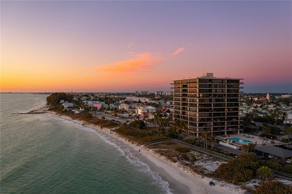 An aerial view shows a tall beachfront condominium building next to a sandy coastline at sunset — 7650 BAYSHORE DRIVE UNIT 401, TREASURE ISLAND, FL