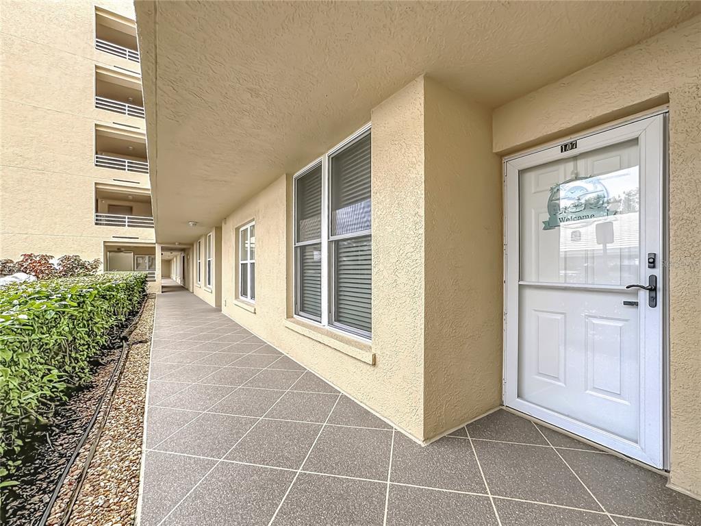 A tiled outdoor walkway leads to a white front door labeled 107 on a building — 3750 PINEBROOK CIRCLE UNIT 107, BRADENTON, FL