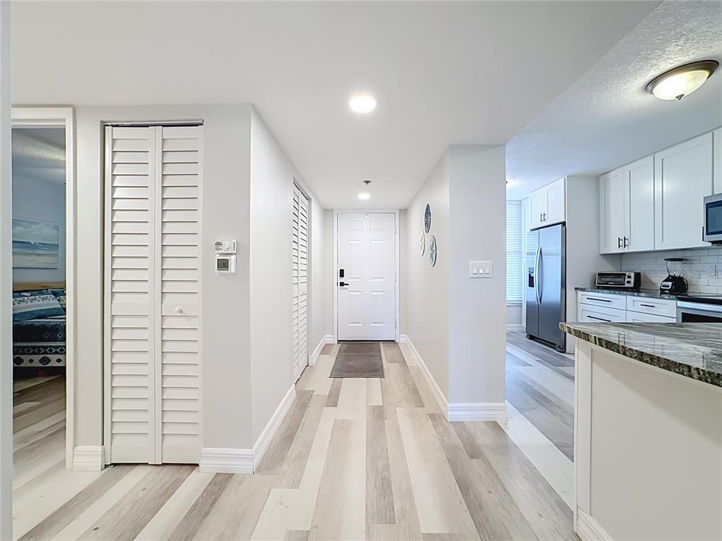 A hallway with light-colored wood-look flooring leads to a white front door and kitchen — 3750 PINEBROOK CIRCLE UNIT 107, BRADENTON, FL