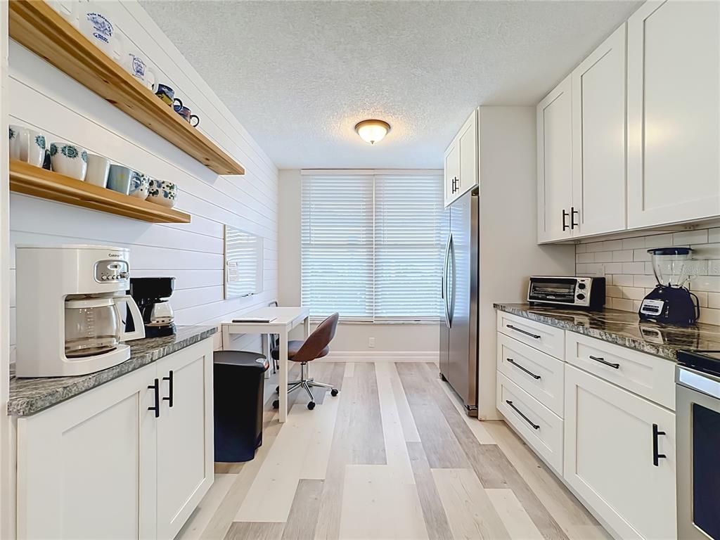 A kitchen features white cabinetry, light wood-look flooring, a small desk area, and open shelving — 3750 PINEBROOK CIRCLE UNIT 107, BRADENTON, FL