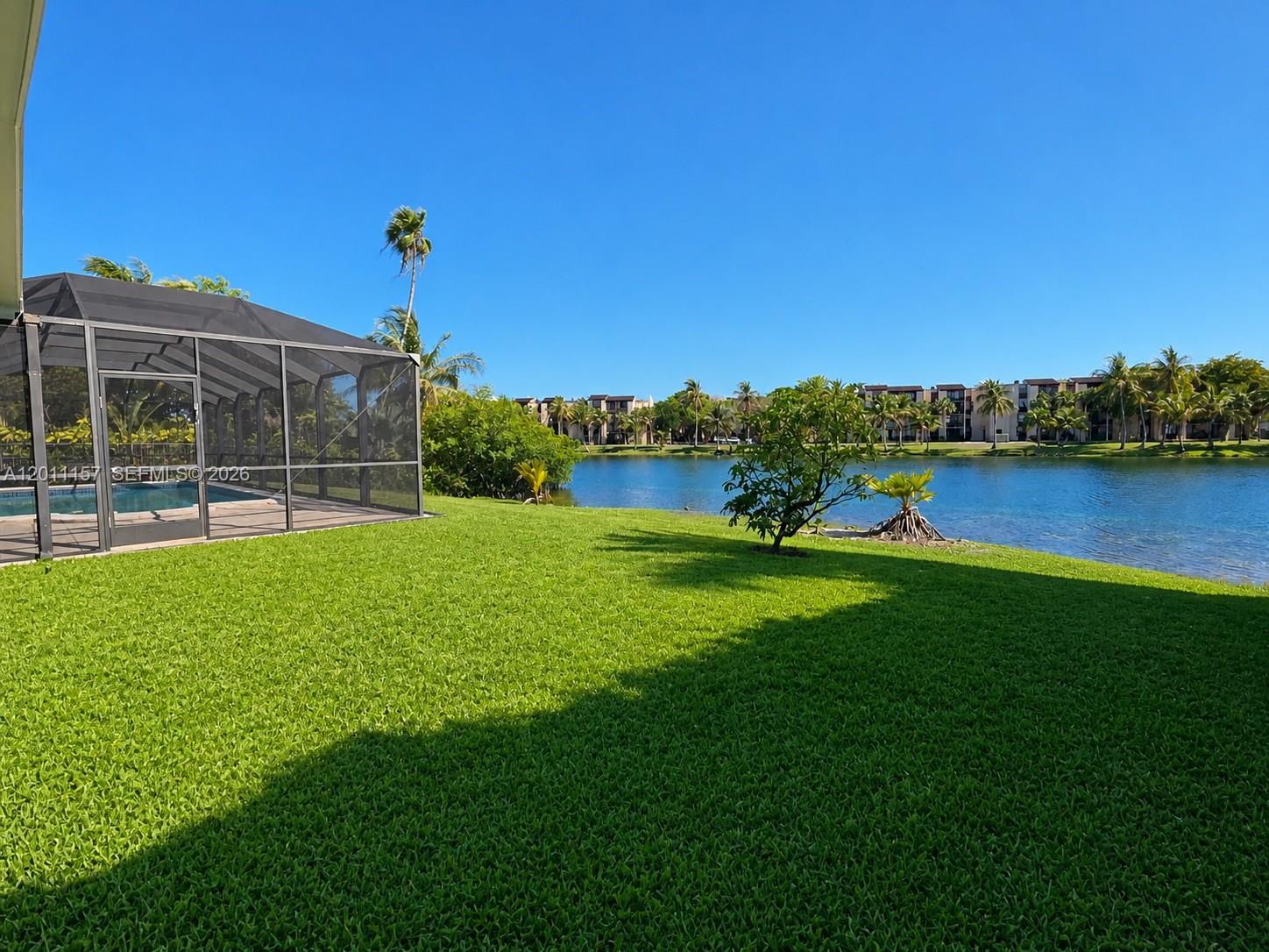 A screened-in pool enclosure sits on a grassy lawn overlooking a lake and palm trees — 14250 SW 85th St, Miami FL 33183, MIAMI, FL