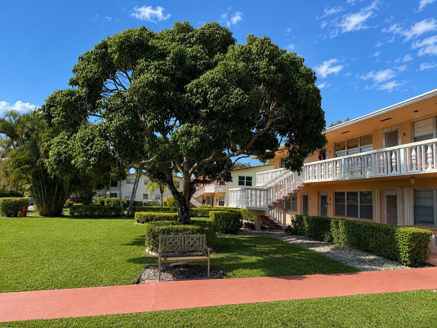 A wooden bench sits under a large tree in front of a two-story apartment building — 67 Camden C, West Palm Beach FL 33417, WEST PALM BEACH, FL