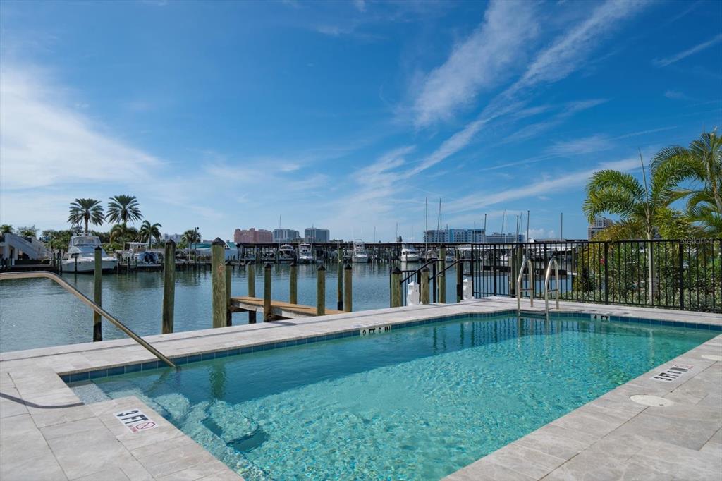 A rectangular swimming pool overlooks a marina with docked boats under a blue sky — 211 DOLPHIN POINT UNIT 501, CLEARWATER, FL