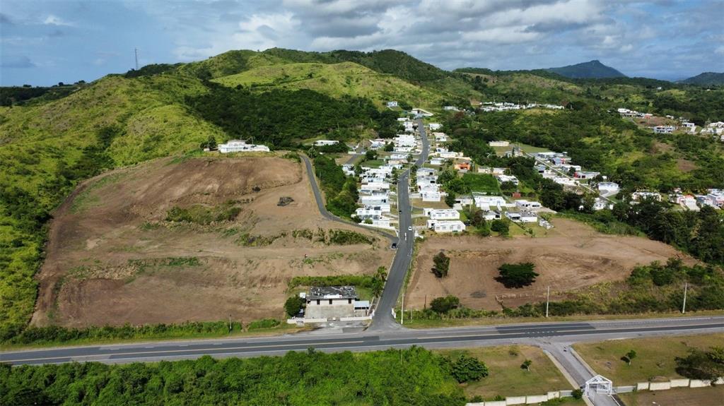 An aerial view shows a paved road leading into a residential neighborhood surrounded by hills — BO. SAN IDELFONSO, COAMO, PR