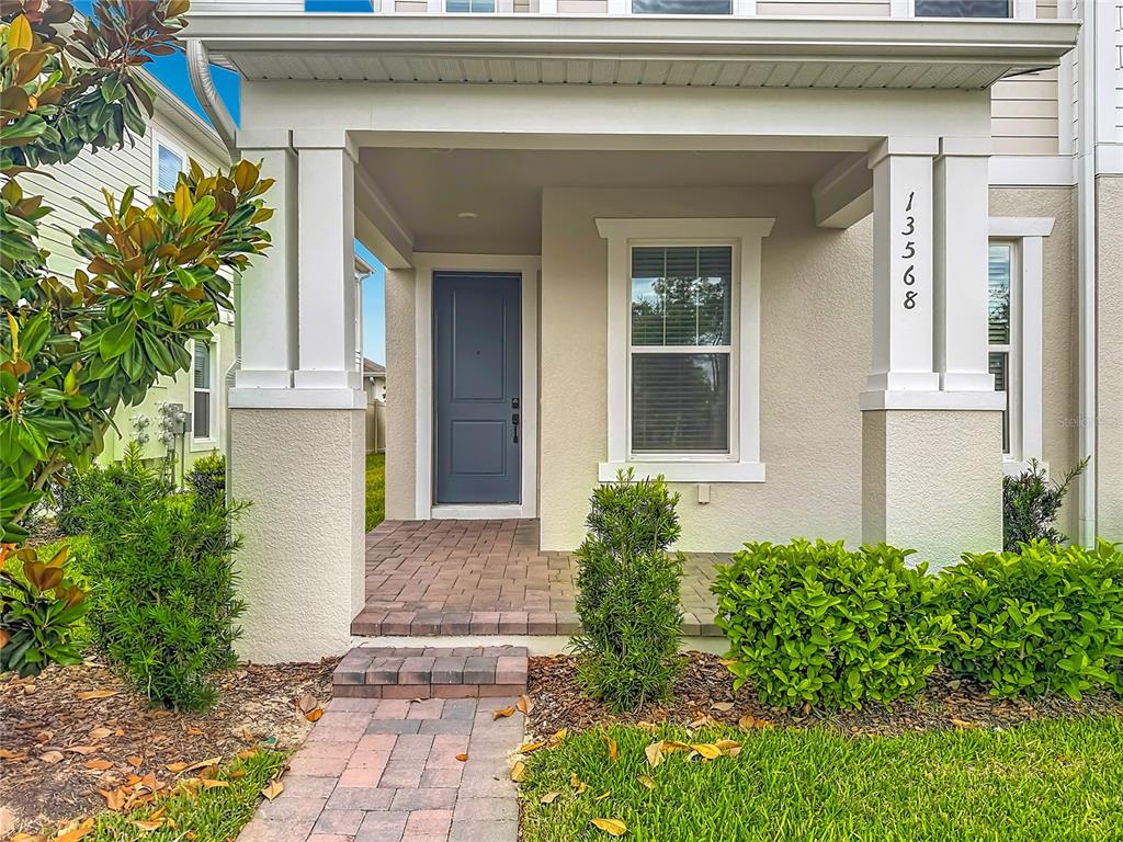A covered front porch with a gray door and brick walkway leads to a house entrance — 13568 AUTUMN HARVEST AVENUE, WINTER GARDEN, FL