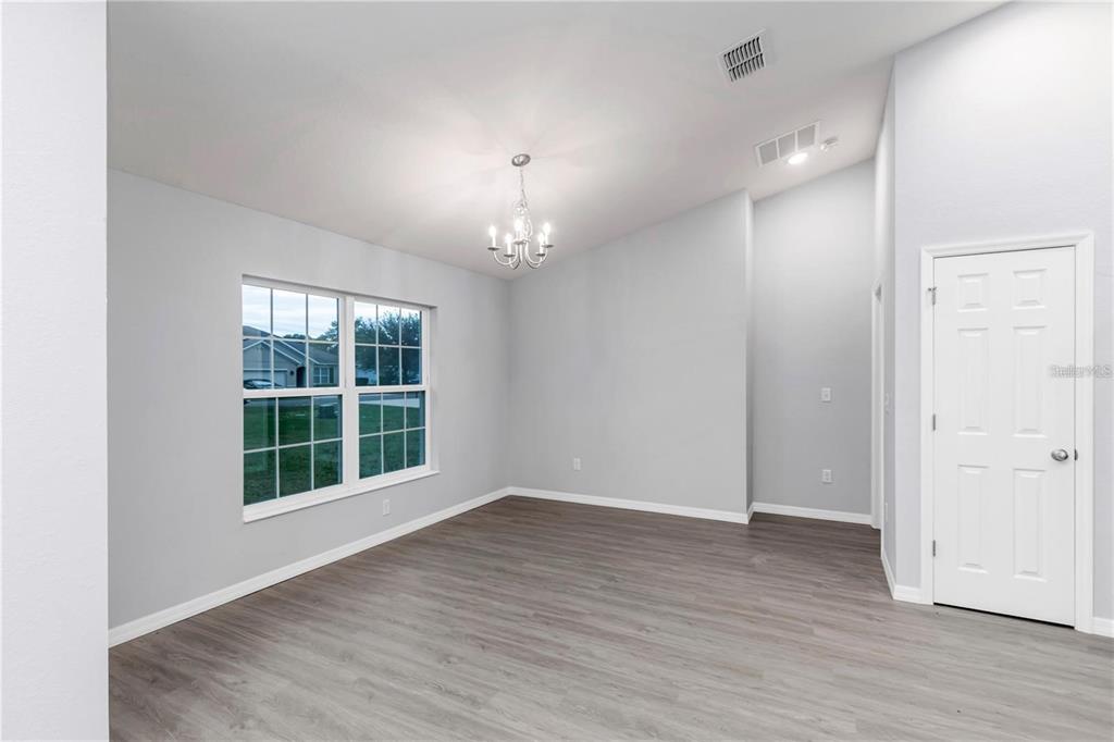 A bright, empty room features light gray walls, wood-look flooring, a chandelier, and a window — 6711 SW 64TH AVENUE, OCALA, FL
