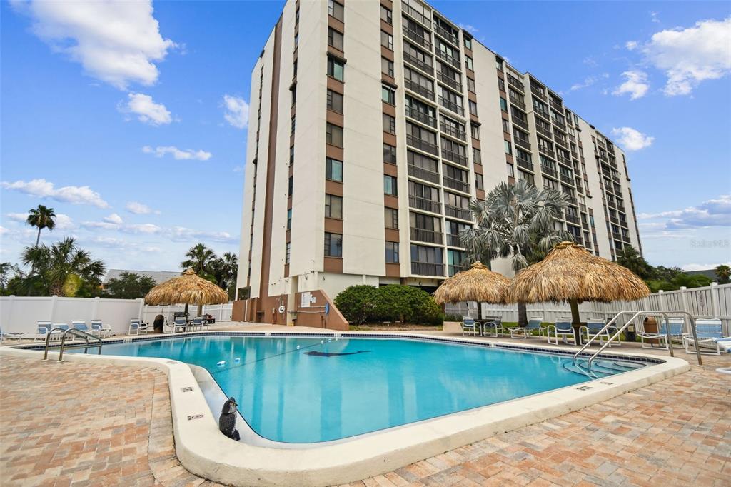A tall apartment building stands behind a swimming pool with several thatched-roof umbrellas — 255 DOLPHIN POINT UNIT 207, CLEARWATER BEACH, FL