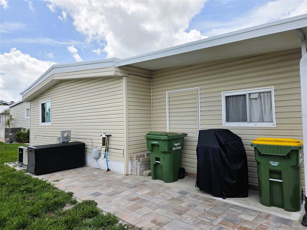 A paved patio area sits beside a beige-sided home with two trash bins and a grill — 3227 BAYBERRY TERRACE, SARASOTA, FL