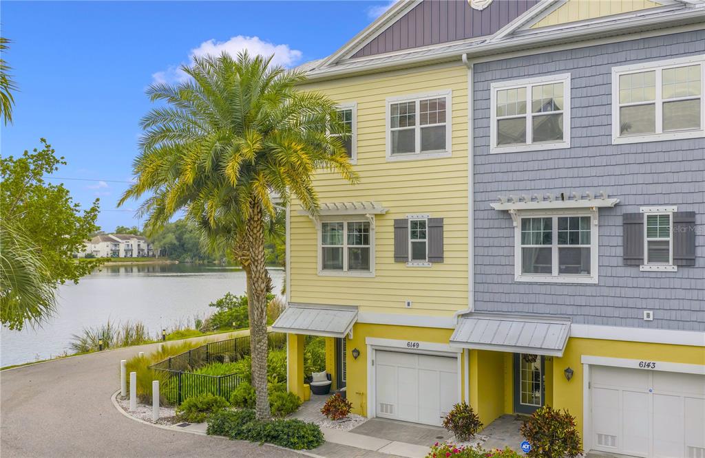 A three-story yellow and gray townhouse stands next to a lake with a palm tree — 6149 ANCHORAGE WAY S, ST PETERSBURG, FL