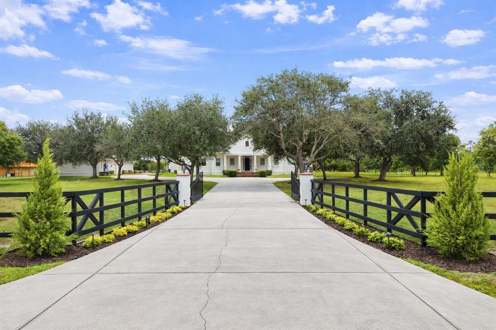 A concrete driveway leads through a gated entrance toward a white house with large trees — 12201 CAISSON LANE, FORT MYERS, FL