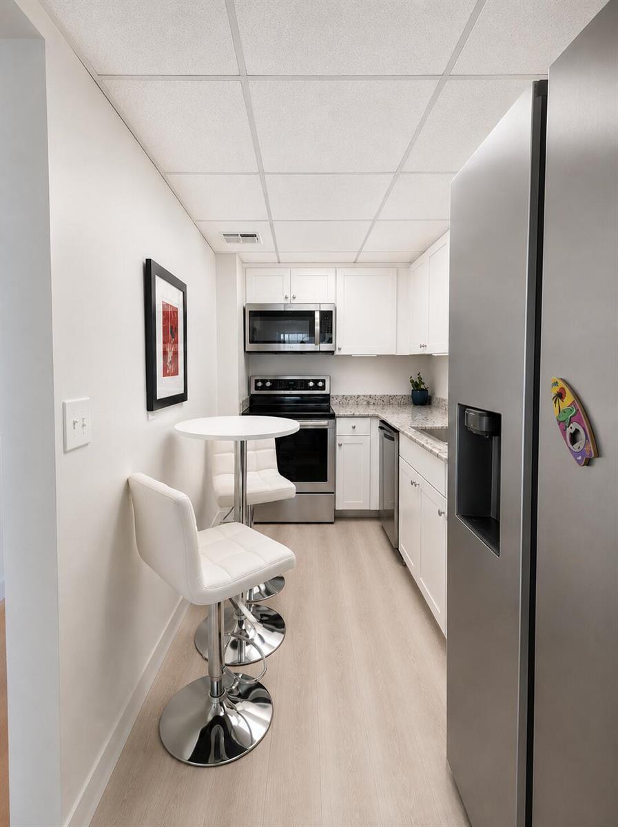 A narrow kitchen features white cabinets, stainless steel appliances, light wood flooring, and a bistro table — 2016 S Federal Highway # 105, Boynton Beach FL 33435, BOYNTON BEACH, FL